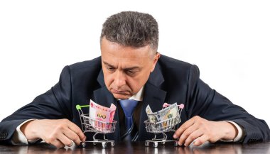 Orta yaşlı takım elbiseli bir adam ve 100 porselen yuan ve beyaz arka planda 100 dolarlık minyatür alışveriş arabaları.