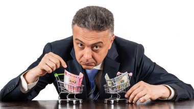 Orta yaşlı takım elbiseli bir adam ve 100 porselen yuan ve beyaz arka planda 100 dolarlık minyatür alışveriş arabaları.