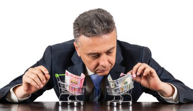 Orta yaşlı takım elbiseli bir adam ve 100 porselen yuan ve beyaz arka planda 100 dolarlık minyatür alışveriş arabaları.