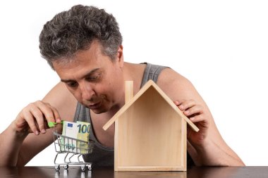 Kolsuz tişörtlü, minyatür ahşap bir ev ve 100 Euro 'luk banknot giyen orta yaşlı fakir bir adam.