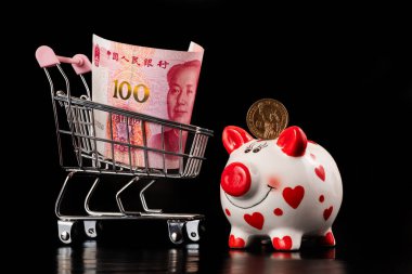 1 Amerikan Doları, domuz kumbarası ve 100 Çin Yuan 'lık minyatür süpermarket alışveriş arabası ve siyah arka plan.