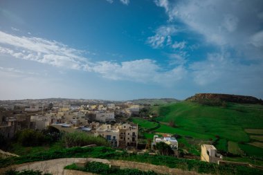 Mavi gökyüzüyle güneşli bir günde Malta 'nın Gozo adasındaki Victoria Kalesi' nden manzara. Etrafta bir sürü yeşil tepe ve ev var..