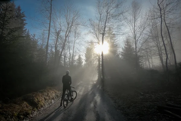 Person resting on a mountain bike ride uphill reaching sun piercing through winter fog or cloudy weather. Winter escape from fog into sun with bike.