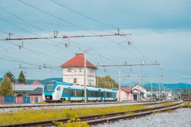 Beyaz ve mavi renkli yolcu treni bir yaz günü Ljubljana Vizmarje istasyonunda hızla ilerliyor. Düşük profilli fotoğraf, istasyondaki diğer trenler görünür.