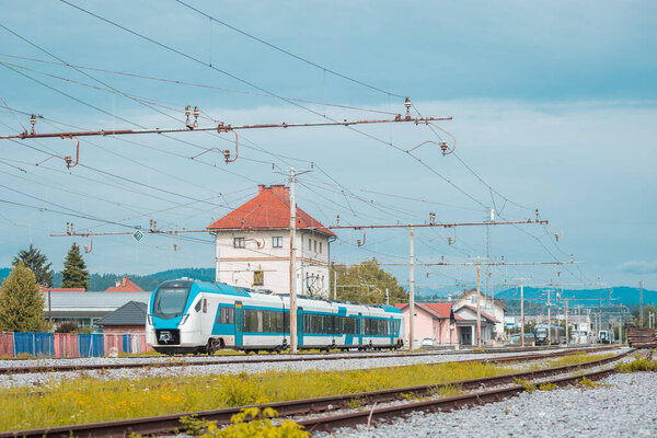 Пассажирский поезд в белом и синем курсирует по станции Любляна Визмарье в летний день. Снимок низкого профиля, другие поезда видны на станции