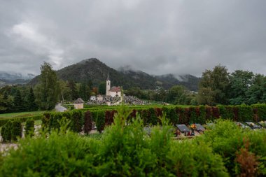 Kamnik kentindeki mezarlık ve ana kilise, önde yeşil yeşillikler, arkada ise kale tepesi bulunuyor. Bulutlu bulanık ayar.