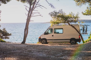 Vanlife 'ı yaşamak. Sahile park etmiş beyaz minibüs karavanı, güzel ağaçlar ve arkadaki mavi gökyüzü. Minibüslü modern göçebe yaşam tarzı.