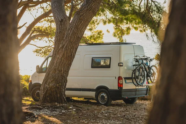 Living the vanlife. White van camper parked on the beach, nice trees around and blue sky just behind the van in the background. Modern nomad lifestyle with a van.