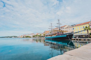 Sonbaharın güneşli bir gününde, Hırvatistan 'ın Losinj adasındaki Mali Losinj kentinin arka planında demirlemiş yelkenli, palmiyeler ve diğer ağaçlarla dolu bir gezinti alanı. Neredeyse hiç insan görünmüyor..