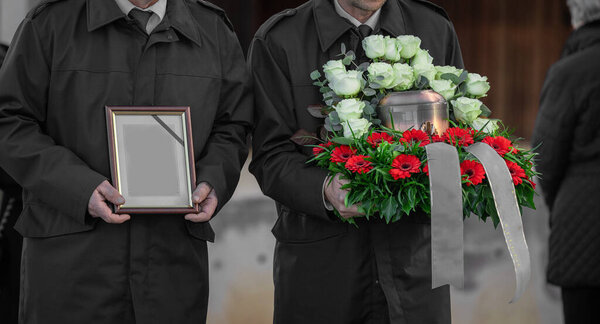Metal urn or funeral container with ash of a deceased person at a memorial service. Undertakers seen carrying the picture and an urn on a last path towards the grave