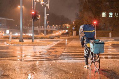 Gıda teslimatı bisikletçisi gece vakti ve yağmur malları dağıtırken, mobil şemsiyeyle korunuyor veya havaya karşı koruyucu kılıfla korunuyor. Kırmızı ışıkta duruyorum..