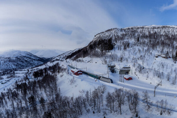 Воздушный беспилотник с видом на норвежский пассажирский поезд, стоящий на платформе на станции Мьёльфьель посреди эпического пейзажа сельской местности. Мбаппе: Эпическая зима.