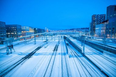Oslo 'daki tren istasyonu otobüs durağındaki üstgeçitlerden birinden görülmüş. Birçok iz istasyona girerken görülüyor, yüksek binalarla çevrili. Akşam mavi saati.