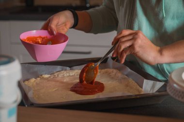 Kadın tavaya yayılmış pizza hamuruna ev yapımı domates sosu döküyor. Ev yapımı pizza evde pişmeye hazırlanıyor.