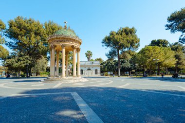 Lecce 'deki büyük bir parkın merkez kısmı. Ortasında sütunlar olan yuvarlak bir anıt. Yeşil ağaçlar ve açık mavi gökyüzü.