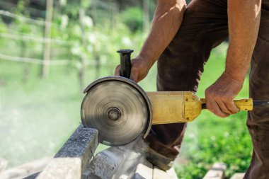 Ev bahçesindeki beton direklerini kesmek için büyük sarı öğütücü kullanan biri. Betonu kesmek için dönen metal bir disk. Görünür döner bıçak. Etrafta uçuşan toz ve enkaz.