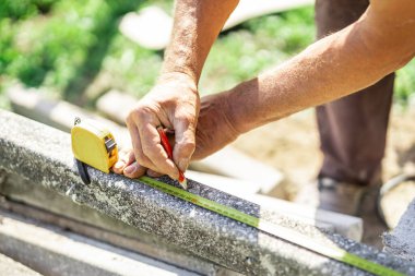 Beton direklerin kesilmeye hazır olduğunu ölçmek için sarı şerit kullanan kişi. Bir elinde kalem, diğer elinde mezura. Bahçede yeşil güneşli bir ortam