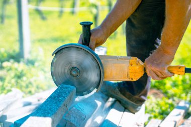 Ev bahçesindeki beton direklerini kesmek için büyük sarı öğütücü kullanan biri. Betonu kesmek için dönen metal bir disk. Görünür döner bıçak. Etrafta uçuşan toz ve enkaz.