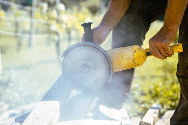 Ev bahçesindeki beton direklerini kesmek için büyük sarı öğütücü kullanan biri. Betonu kesmek için dönen metal bir disk. Görünür döner bıçak. Etrafta uçuşan toz ve enkaz.