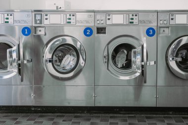 View of a modern stainless steel machines in a laundromat or laundrette. Public laundry with multiople washing machines. Frontal view, one door open clipart