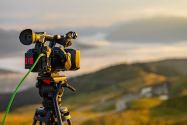 Modern video kamera bir tripodun üzerinde çekim yapıyor. Güzel bir sabah panoraması. Sabahları bir kameraman için tipik bir iş. Arka tarafında Sdi sinyal kablosu olan el kamerası.