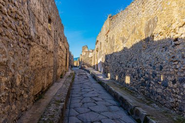 Pompei arkeoloji sahasındaki kaldırım taşlı Roma yollarından biri. Duvarları ve aralarında yol olan görünür kalıntılar. Güneşli bir gün, arka planda Vezüv Dağı...