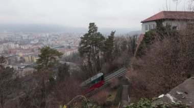 Graz iz 'de kırmızı renkli modern füniküler üst istasyona tırmanıyor. Arka planda Graz şehrinin büyük manzarası, bulutlu kış günü.