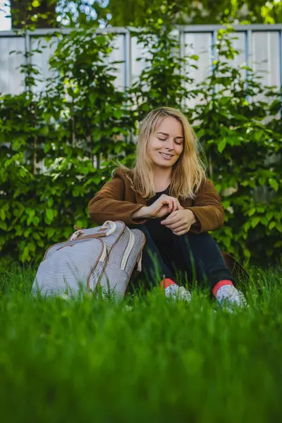 Sırt çantalı, kahverengi ceketli, siyah pantolonlu, çimenli bir çimenlikte oturan genç bir kadın ya bahçedir ya da çalılarla kaplı bir parktır. Görüntünün genel havası huzurlu ve rahat..