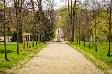 Tivoli Parkı, Ljubljana 'da, çıplak ağaçlar ve yeşil çimlerle kaplı çakıllı bir patika, tarihi bir bina ve arka planda daha fazla yeşillik bulunan bir heykele öncülük eder.