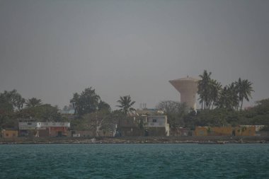 Saint-Louis, Senegal 'deki rıhtım kenar mahalleleri, mütevazı evleri, palmiye ağaçları ve su kulesi olan dağınık bir kıyı şeridi gösteriyor. Enkaz, sakin ve yansıtıcı suyun üzerinde puslu bir gökyüzü ile kenarı kirletir.