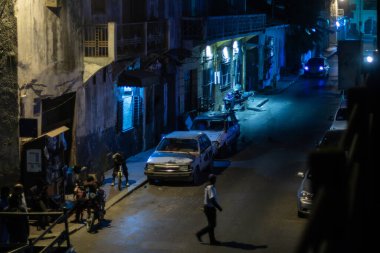 Saint-Louis, Senegal 'de bir gece sokak sahnesi, eski binalar, park edilmiş arabalar, motosikletler ve yürüyen insanlar. Mavi sokak lambalarıyla aydınlatılan bu ışık, canlı ama kasvetli şehir atmosferini yakalıyor..