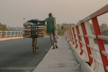 Biri Ziguinchor, Senegal 'deki Casamance Nehri' ni kapsayan bir köprüye tahta kalaslarla dolu bir bisiklet itiyor. Sahnede kırmızı-beyaz bir korkuluk, sisli bir gökyüzü ve günlük yaşamı ve labirenti gözler önüne seren uzak ağaçlar yer alıyor.