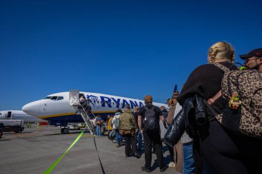 Berlin, Almanya, 28.4.2025: Yolcular açık mavi bir gökyüzünün altındaki pistte seyyar bir merdivenden Ryanair uçağına biniyor. Uçak mavi ve sarı üniformalı havayollarına sahiptir. Yer ekibi ve ekipmanlar görülebiliyor, gezginler çanta taşıyor.