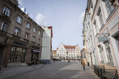 Tallinn 'in eski kasabasındaki bir kaldırım taşı sokakta dükkanları ve kafeleri olan tarihi binalar yer alıyor. İnsanlar, şehrin ortaçağ cazibesini sergileyen, parçalı bulutlu bir gökyüzünün altında, kapalı bir binayla şehir meydanına doğru yürüyorlar..