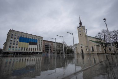 Yağmurlu Tallinn caddesi yeşil çatılı bir kiliseyi ve Estonya bayrağını gösteren modern bir binayı yansıtır. Bulutlu gökyüzü ve ıslak kaldırım ağaçlar ve sokak lambalarıyla kasvetli bir şehir manzarası yaratır...