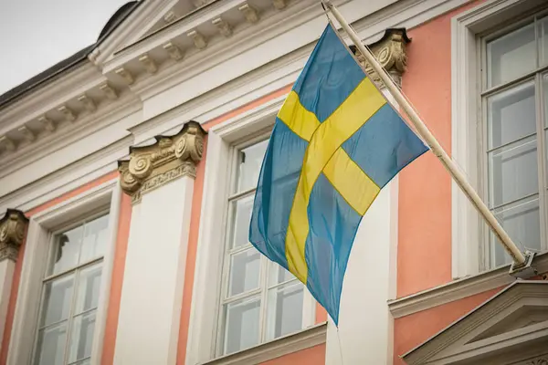 Bir İsveç bayrağı Tallinn 'deki tarihi bir binanın önünde dalgalanıyor, şeftali rengi duvarlar, beyaz süslemeler ve bulutlu bir gökyüzünün altında süslü detaylar yer alıyor,.