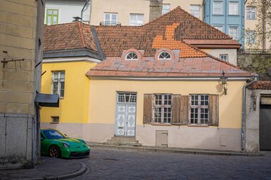 Parlak yeşil bir spor araba Tallinn 'in Eski Kasabası' nda bir kaldırım taşı caddesinde duruyor, eskimiş panjurları ve kırmızı fayanslı çatısı olan büyüleyici sarı bir evin yanında, tarihi ve modernliği harmanlayan.