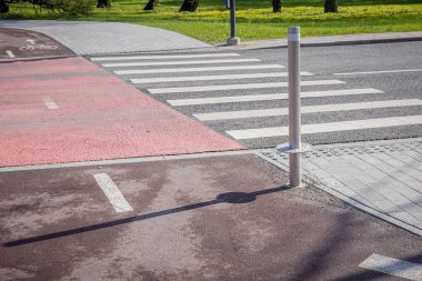 Kırmızı bisiklet şeridi, modern bir şehir ortamında bir zebra geçidinden geçiyor. Sahnede gölgesi olan metal bir direk, bitişik bir kaldırım ve arka planda ağaçlar olan yeşil bir park yer alıyor. Yol ıslak, gün ışığını yansıtıyor, temiz şeritli.