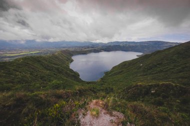 Görüntü Cuicocha Gölü 'nü gösteriyor. Ekvador' da, volkanik bir kalderanın içine yerleştirilmiş çarpıcı bir krater gölü. Derin mavi sular, sarp, yeşil tepeler ve bulutlu bir gökyüzünün altındaki uzak dağlarla çevrili gökyüzünü yansıtır.