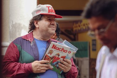 Quito, Ekvador, 11.2.2017: Kırmızı ve beyaz Tako şapkalı ve renkli ceketli bir kişi açık hava ortamında bir yığın Nacho Libre kitabı tutuyor. Arka plan bulanık, başka biri ve sarı bir duvar görünüyor. Bu da canlı bir sokak manzarası olduğunu gösteriyor.