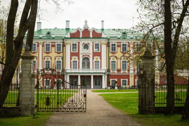 Tallinn 'deki büyük şatoda kırmızı, sarı ve beyaz aksanlı Barok tarzı bir ön cephe, yeşil çatı ve süslü detaylar var. Çakıl yolu, taştan sütunlarla çevrili, açık, dekoratif demir bir kapıya götürür, yemyeşil çimenler ve ağaçlar çerçevelenir.