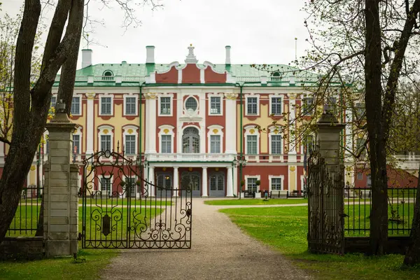 Tallinn 'deki büyük şatoda kırmızı, sarı ve beyaz aksanlı Barok tarzı bir ön cephe, yeşil çatı ve süslü detaylar var. Çakıl yolu, taştan sütunlarla çevrili, açık, dekoratif demir bir kapıya götürür, yemyeşil çimenler ve ağaçlar çerçevelenir.