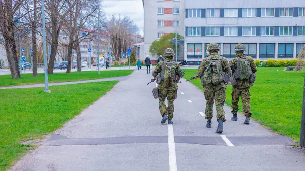 Narva 'da, üç asker kamuflaj devriyesinde otlağın yakınındaki asfalt bir yolda devriye geziyor. Yol boyunca çıplak ağaçlar, çok katlı bir bina ve arka planda bulutlu bir gökyüzünün altındaki diğer kentsel yapılar var..