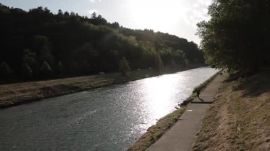 Celje 'deki Savinja Nehri akşam ışığında parlıyor. İnsanlar nehir kenarındaki patika boyunca yürürler, etrafları orman ve sakin doğa ile çevrilidir. Gün batımında Alec 'e nehir kıyısından bak..