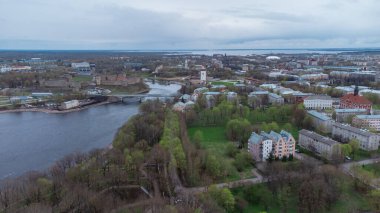 Narva, Estonya, Narva Nehri, yemyeşil ve ağaçlarla çevrili. Ufuk çizgisinde, Hermann Kalesi kalıntıları ve bir kule de dahil olmak üzere çağdaş ve tarihi binaların bir karışımı yer alıyor. Bulutlu gökyüzü sönük bir ışık saçar