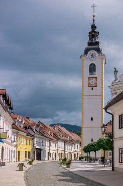 Slovenya 'nın Kamnik kentindeki eski caddede kaldırım taşları, tarihi pastel binalar ve ikonik kilise kulesi bulunuyor. Yerli halk bulutlu bir gökyüzünün altında huzur içinde yürüyor..