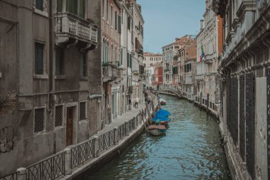 Venedik 'teki dar kanallar yıpranmış duvarları, balkonları ve kemerli pencereleri olan tarihi binalar arasında. Mavi kapaklı küçük tekneler su üzerinde yüzer ve kanal üzerinde küçük bir köprü kemerleri vardır. Çok az insan dar patika boyunca yürür.