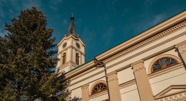 Wis 'te tarihi bir kilisenin parçası olan saati olan klasik bir çan kulesi, açık mavi gökyüzüne karşı uzun bir ağaç tarafından çerçevelenmiş. Kemerli pencereleri ve sütunları olan zarif mimari bu manzaralı kenti vurguluyor..