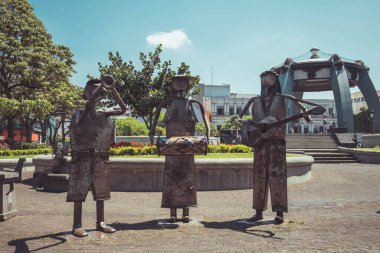 Parkta trompet, davul ve gitar çalan müzisyenlerin üç metal heykeli. Çembersel bir platformda yeşil ve çiçeklerle dururlar, açık mavi bir gökyüzünün altında. Arka planda bir pavyon ve binalar var, bankta oturan bir kişi var.