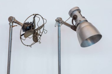 İki eski endüstriyel cihaz: açıkta bırakılmış bıçakları olan bir metal yelpaze ve büyük konik gölgeli sağlam bir lamba, her ikisi de düz beyaz arka plana monte edilerek sade beyaz bir zemine monte edilmiş, kırsal tasarımlarını sergiliyorlar..